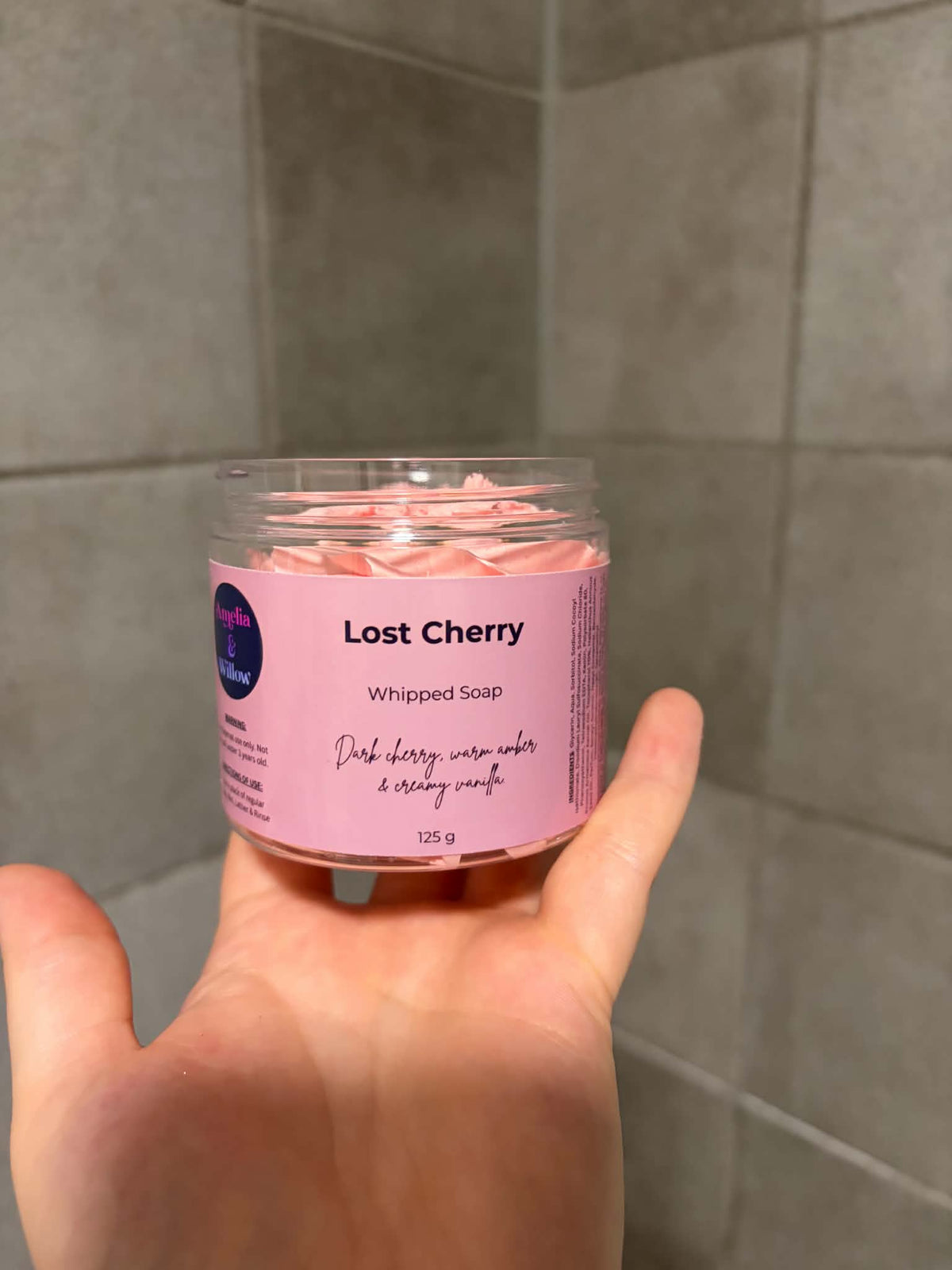 A hand holding a clear plastic jar of Nourish & Natural 'Lost Cherry' whipped soap against a gray tiled background.