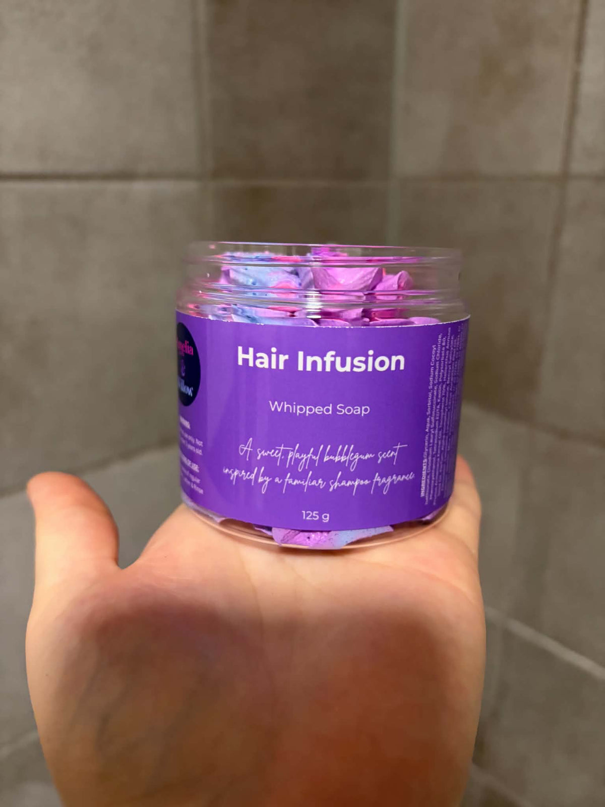 A hand holding a clear plastic jar of Hair Infusion Whipped Soap against a tiled bathroom wall background.
