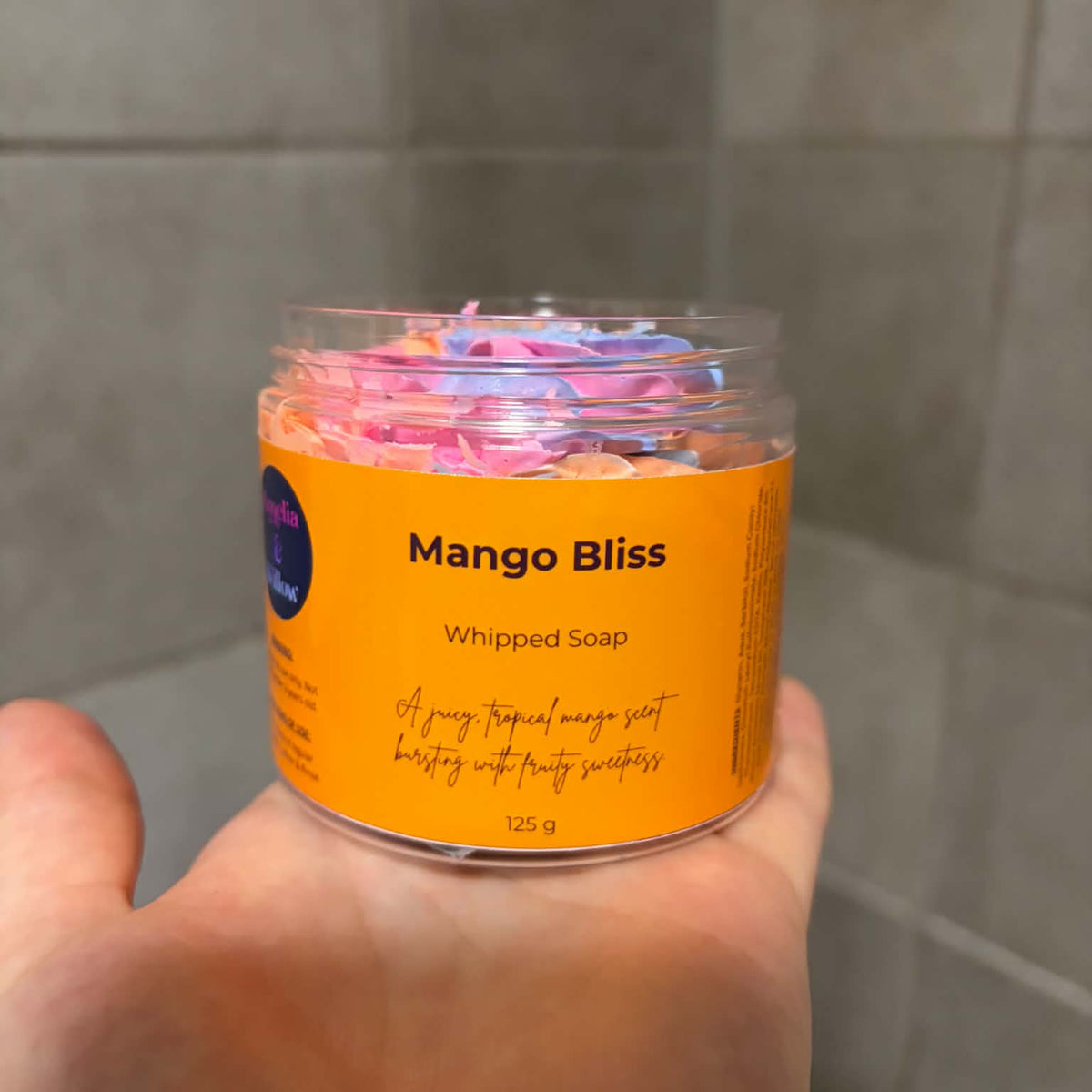 A hand holding a clear plastic jar of Mango Bliss Whipped Soap, showing the colorful whipped soap inside and an orange label, against a gray tiled background.