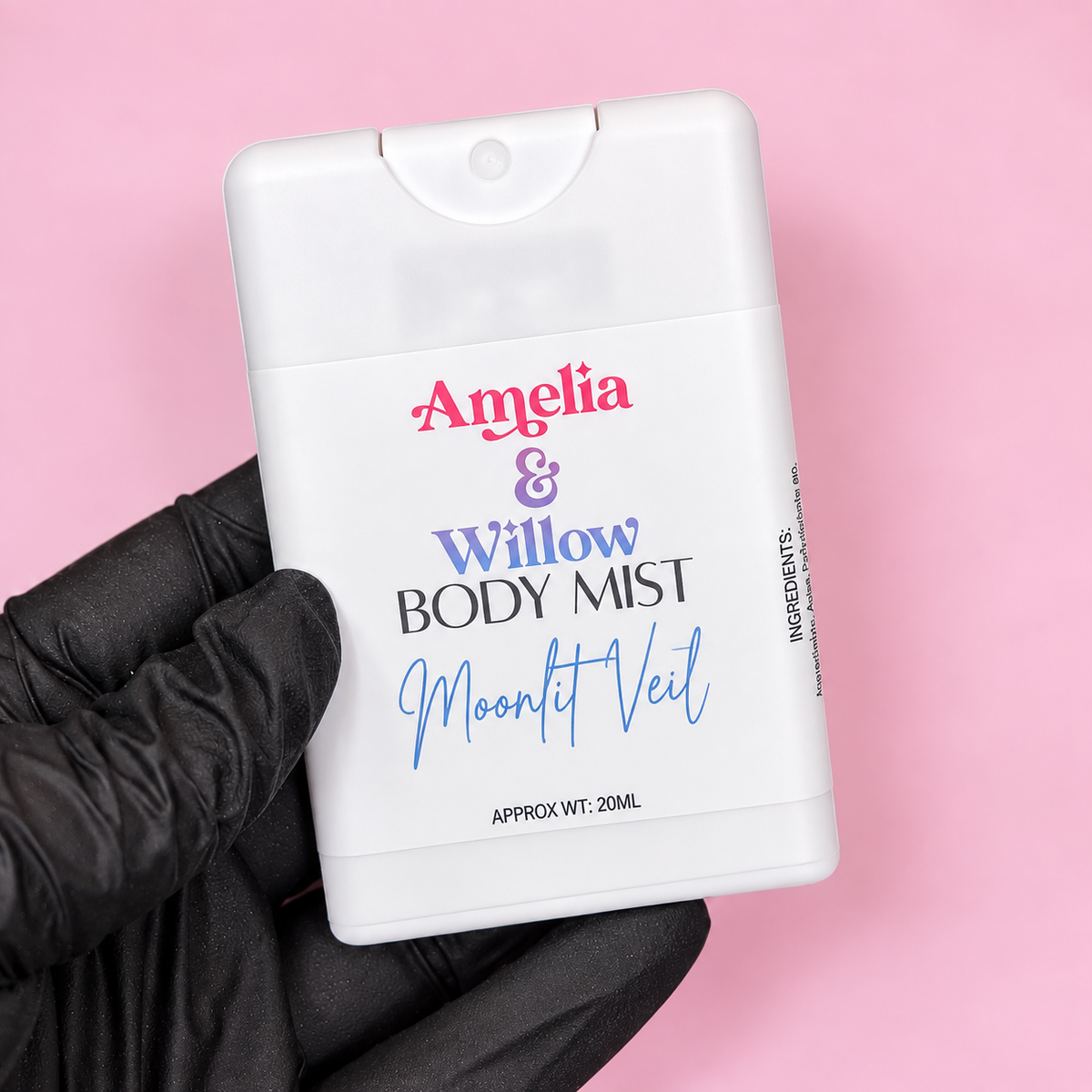 A hand wearing a black glove holds a white plastic body mist bottle against a solid pink background. The bottle is personalized with the names 'Amelia & Willow' in pink and purple, and 'MOONlit Veil' in blue script.