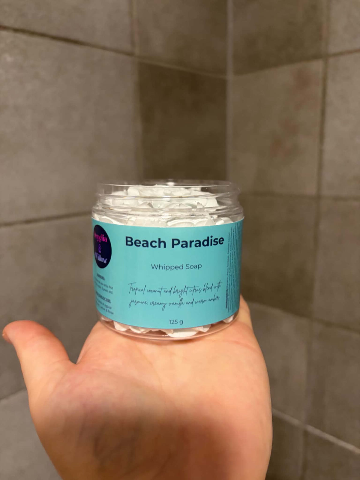 A hand holding a clear plastic jar of Beach Paradise Whipped Soap against a tiled bathroom wall background.

