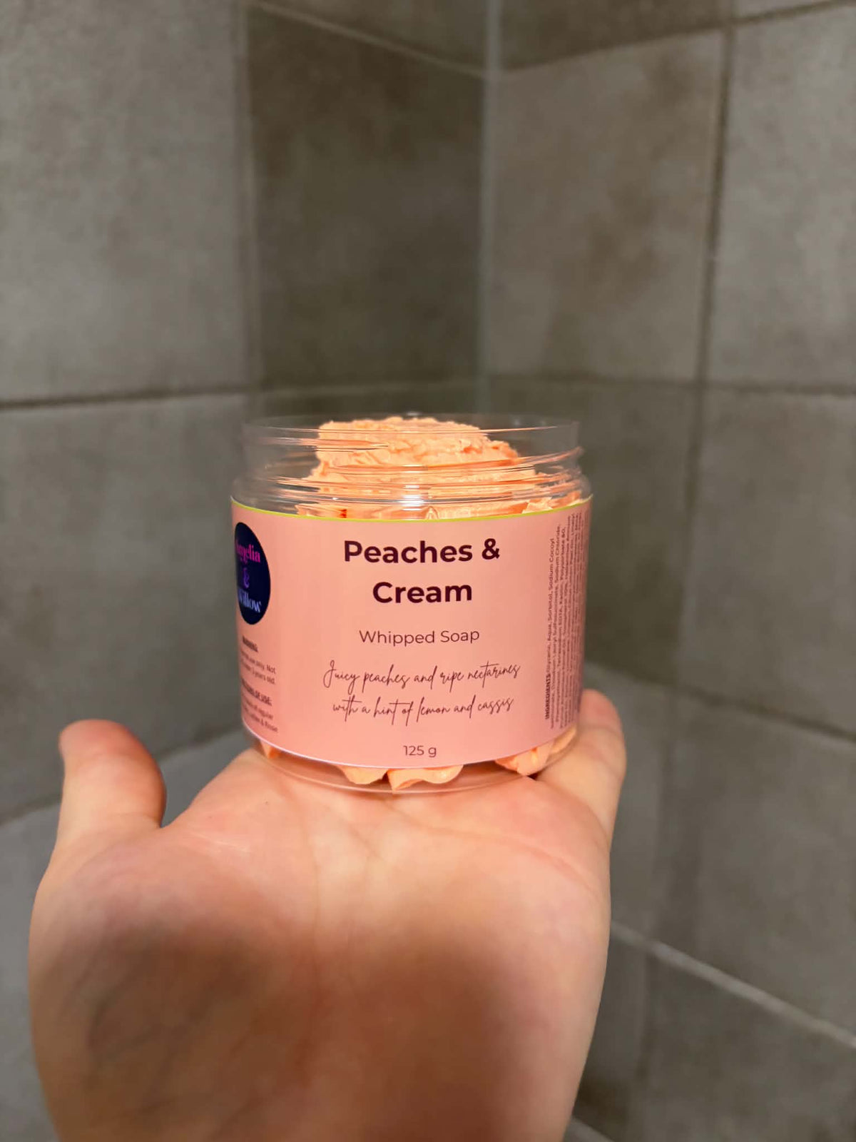 A hand holding a clear plastic jar of Peaches & Cream Whipped Soap against a gray tiled background.

