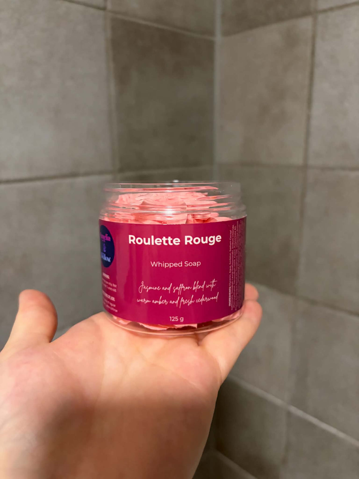 A hand holding a clear plastic jar of Roulette Rouge Whipped Soap against a gray tiled background.

