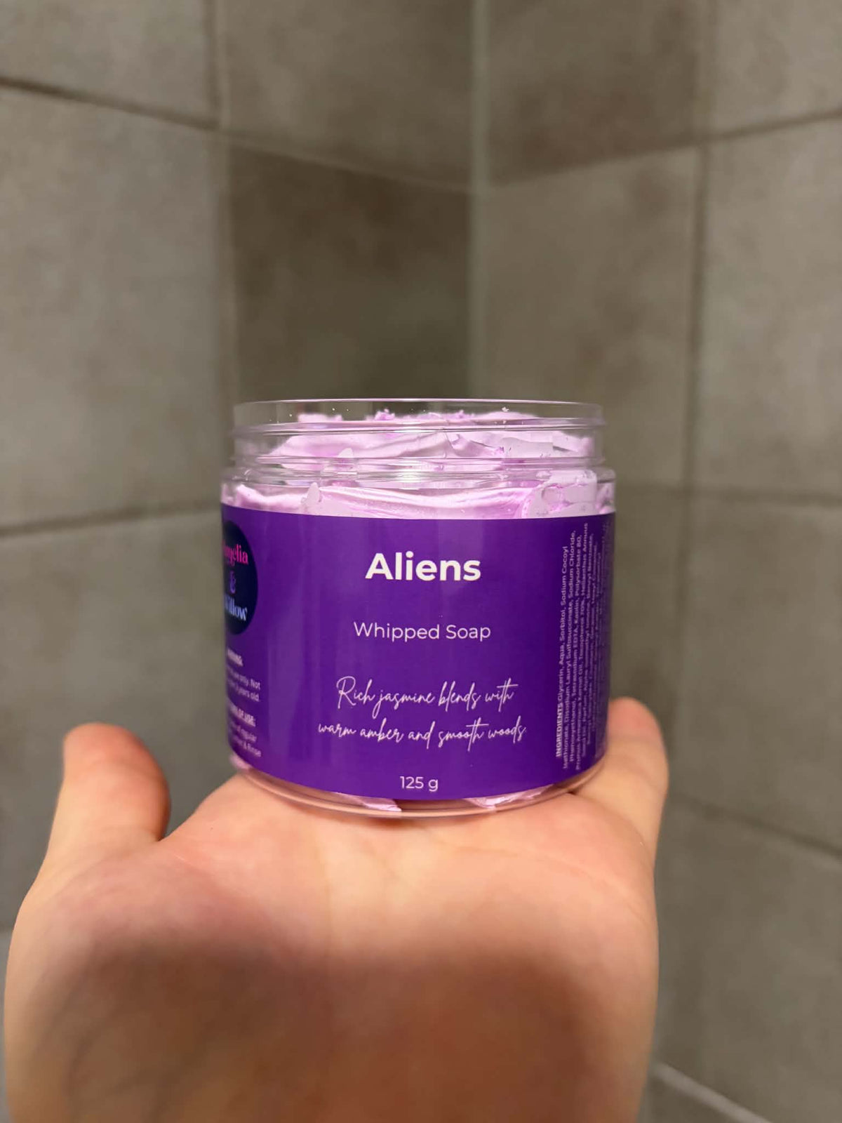 A hand holding a clear plastic jar of Aliens Whipped Soap, showing the light purple whipped product inside, against a tiled gray bathroom wall background.

