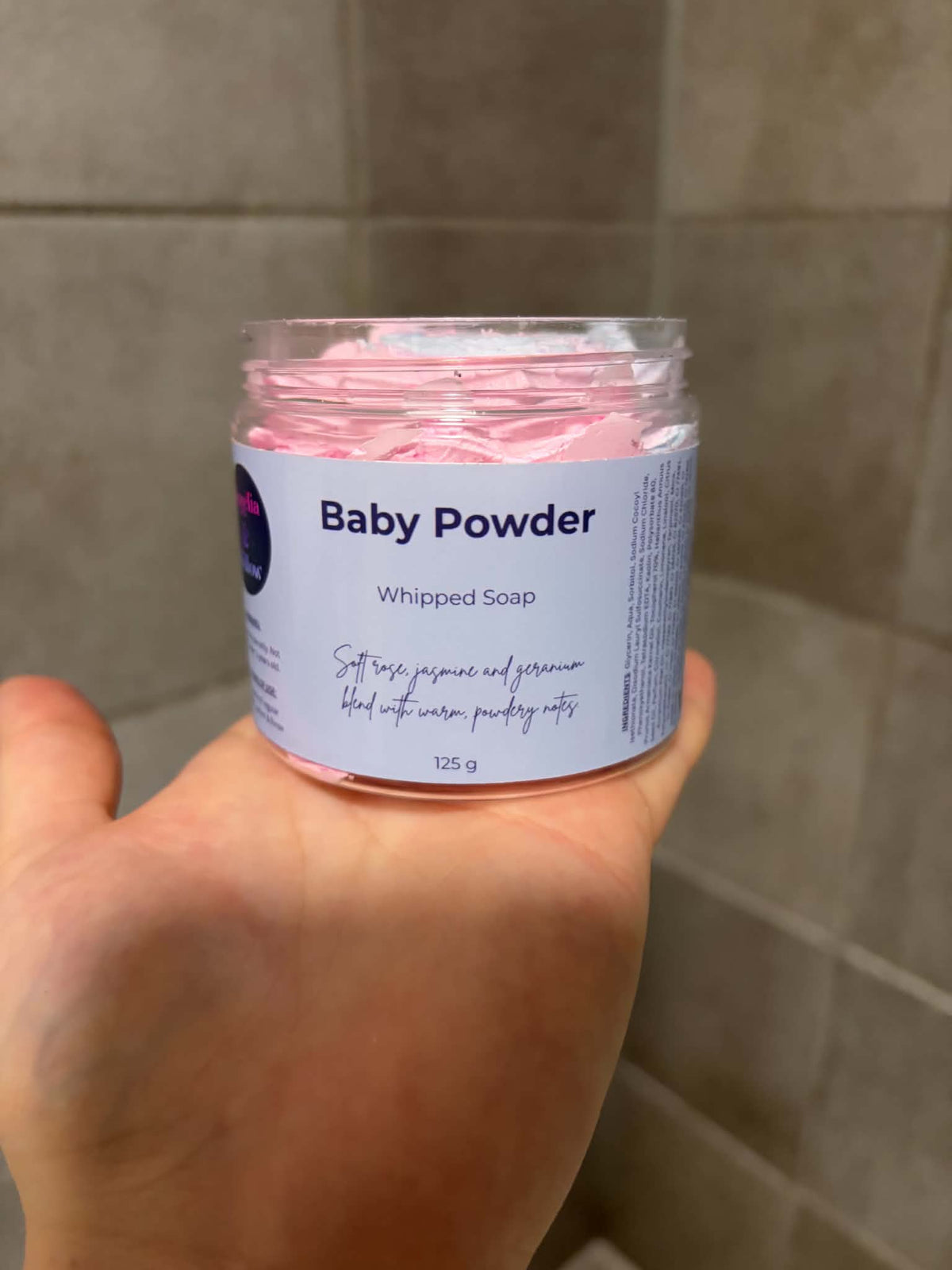 A hand holding a clear plastic jar of Baby Powder Whipped Soap against a tiled bathroom wall background.


