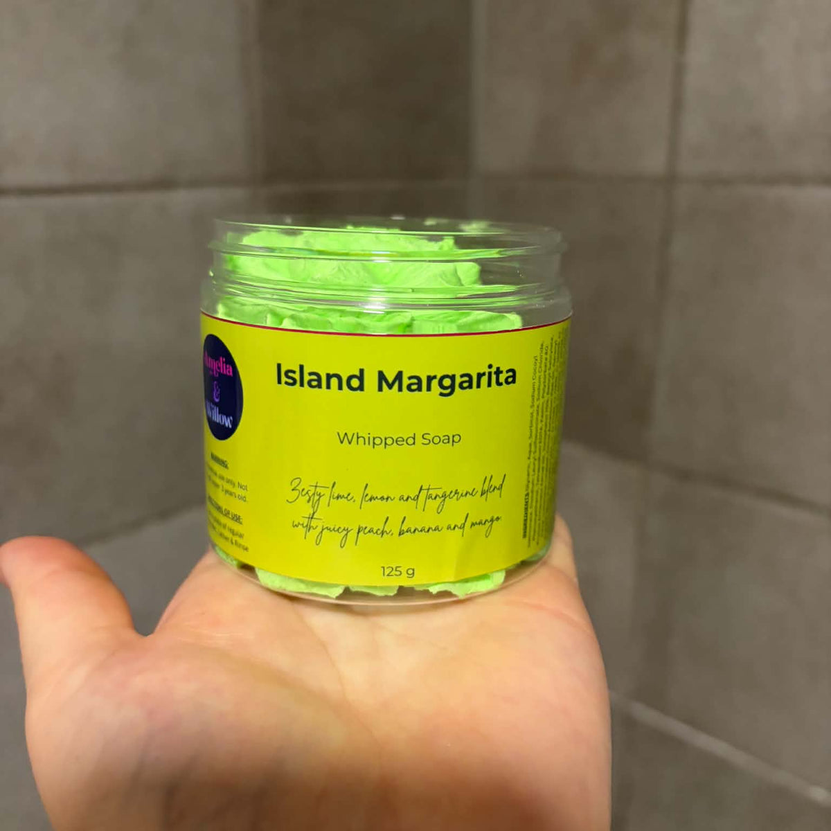 A hand holding a clear plastic jar of whipped soap labeled 'Island Margarita' against a tiled bathroom wall.

