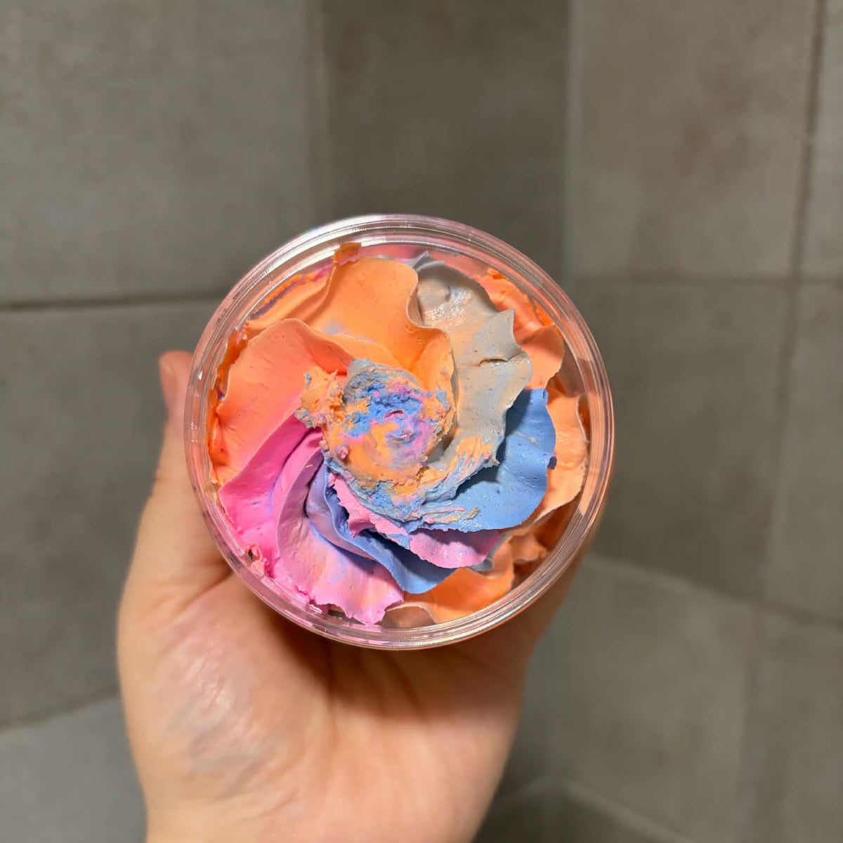 A hand holding a clear round plastic jar filled with a colorful, textured cream or wax product in shades of orange, pink, purple, and blue, set against a gray tiled background.

