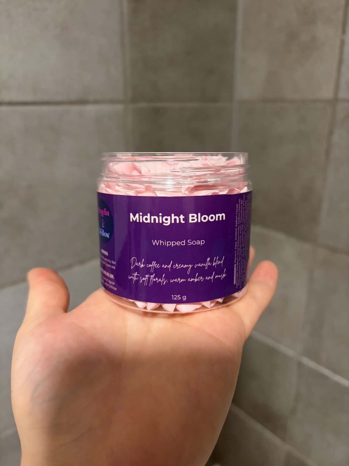 A hand holding a clear plastic jar of whipped soap labeled 'Midnight Bloom' against a gray tiled background.

